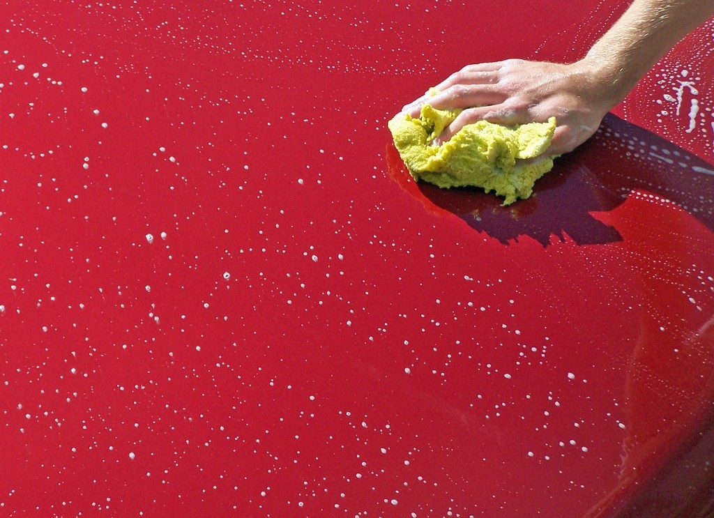 Foto que muestra una mano sosteniendo una esponja, lavando el capó de un auto rojo.
