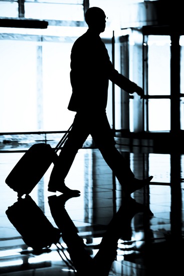Silueta de un hombre con traje caminando en un aeropuerto tirando de una pequeña maleta.