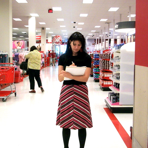 Una mujer sosteniendo y escribiendo en un cuaderno se encuentra dentro de una tienda Target. Otros compradores están en el fondo.