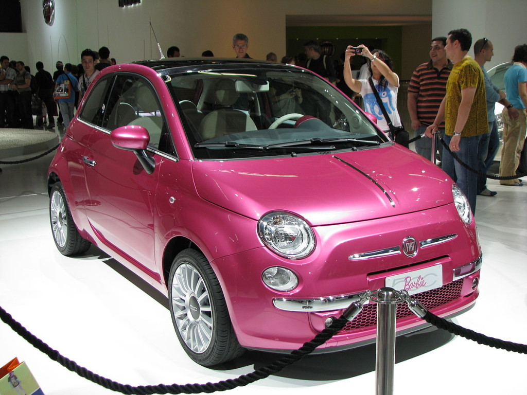 Un auto rosa de dos puertas en un piso de exhibición de autos muy iluminado.
