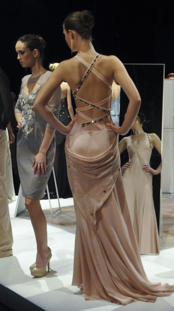 Three runway models wearing gowns pose in front of mirrors.