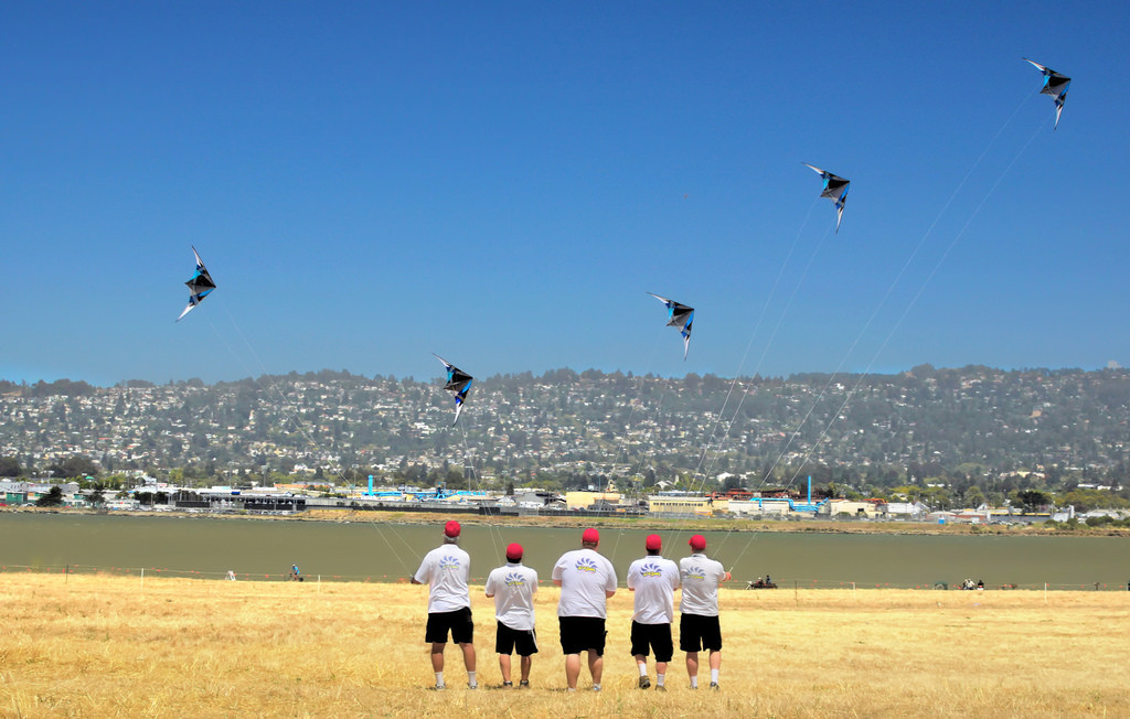 Five men wearing identical hats, shirts, and shorts. They are each flying identical kites.