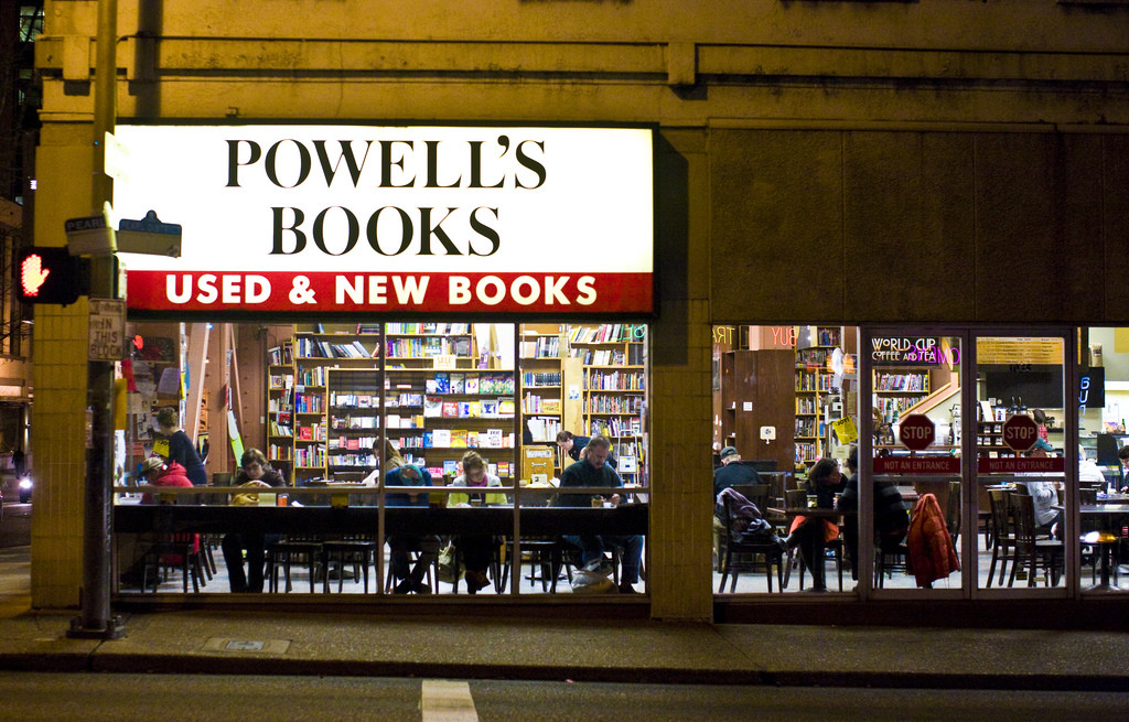 Una tienda iluminada para la librería Powell's Books. Los grandes ventanales muestran estantes altos llenos de libros y mesas donde la gente está sentada, comiendo y leyendo.