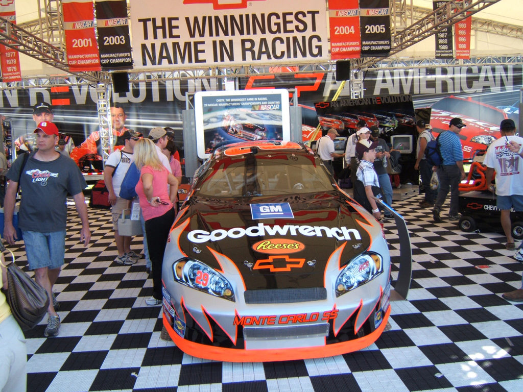 A group of people walking around a Nascar display area. There is a large Chevy racecar parked inside, covered in decals such as G M, Reese's candy, and Good wrench.
