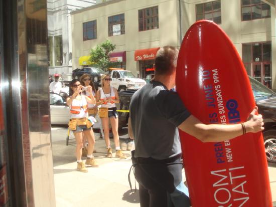 Photograph of a man holding a bright red surfboard. An ad for "John from Cincinnati," an HBO show, is printed on the surfboard.